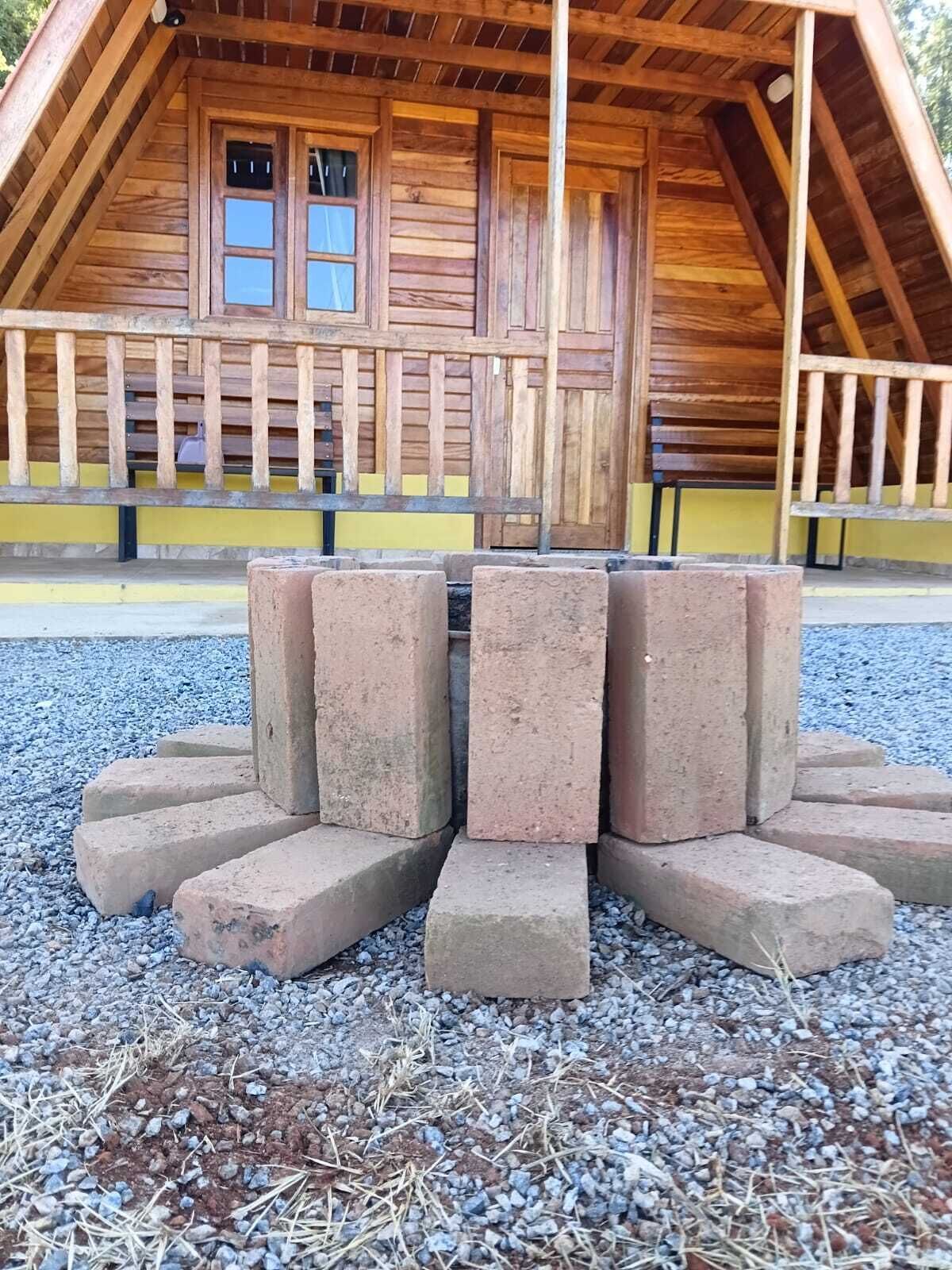 A wooden cabin with a pitched roof, a porch with a railing, and a fire pit made of stacked stones.