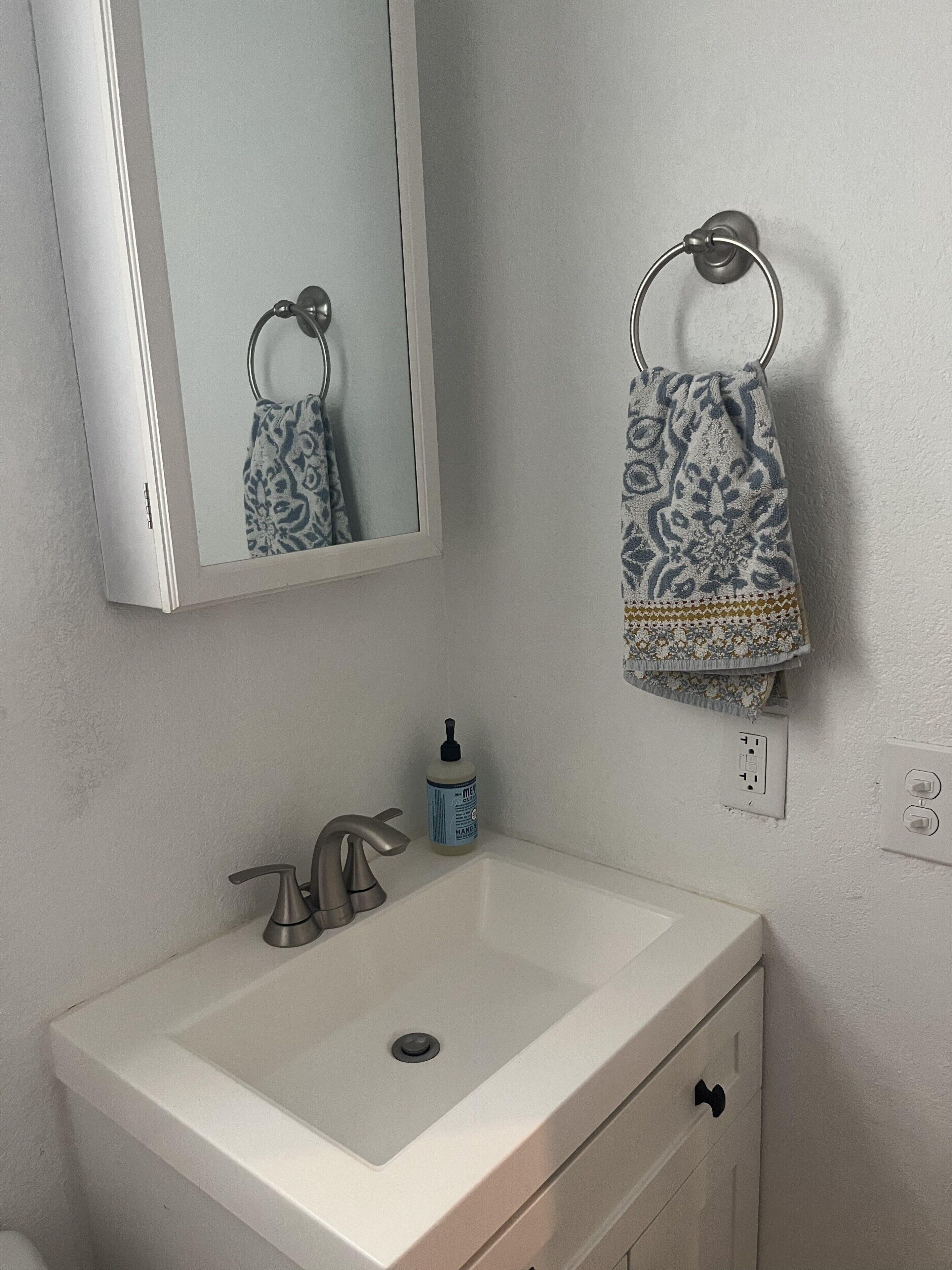 A bathroom with a white sink, a mirror, a towel ring with a patterned towel, and a bottle of hand soap.