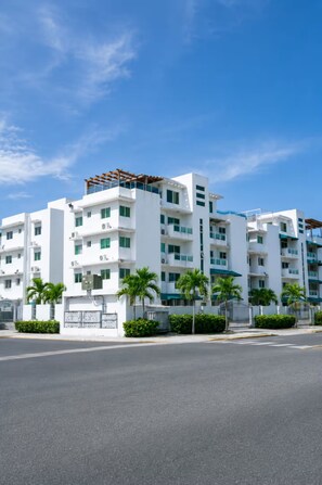 A multi-story hotel with a modern design, featuring balconies, air conditioning units, and a gated entrance.