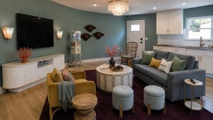 Ein Wohnzimmer mit einer Couch, Hocker, einem Couchtisch, einem Fernseher und einer Küchenzeile mit Schränken und einem Fenster.