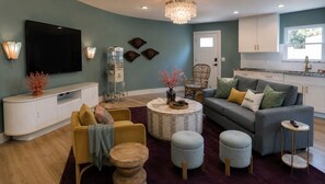 A living room with a sofa, ottomans, a coffee table, a TV, and a kitchen area with cabinets and a window. - Steps to Beach + Private Pool, Theater & Arcade (St. Augustine)