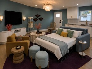 A modern bedroom with a bed, sofa, ottomans, a TV, and a dining area. - Steps to Beach + Private Pool, Theater & Arcade (St. Augustine)