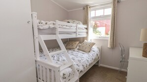 Una cama individual con estructura blanca y ropa de cama floral, una ventana con cortinas y un pequeño escritorio con lámpara.