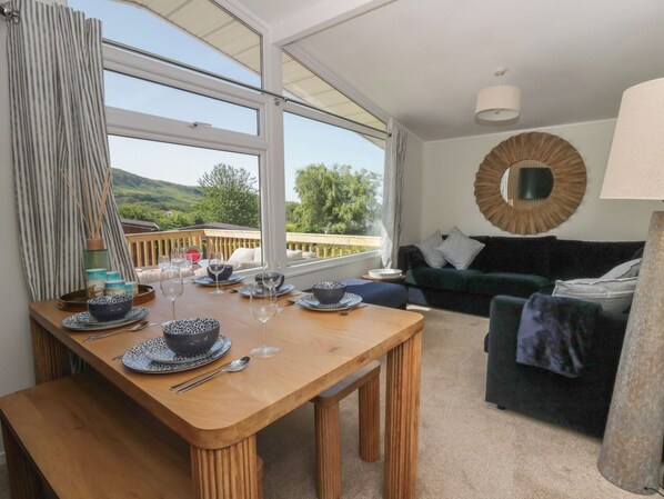 A dining area with a wooden table set for two, a sofa, and a view of the outdoors. - Oakland (Tywyn)