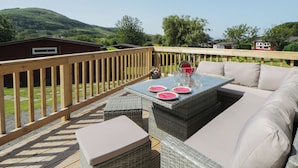 Una terraza al aire libre con un sofá de mimbre, una mesa con vasos y rebanadas de sandía, y una barandilla de madera.