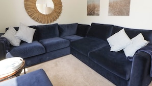 Una sala de estar con un sofá seccional azul oscuro, dos almohadones a juego, un espejo redondo de madera y dos cuadros de paisajes en la pared.