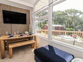 Una habitación con una mesa de madera dispuesta para comer, un banco, un televisor montado en una pared de listones y un ventanal con vistas a un patio cercado y árboles.