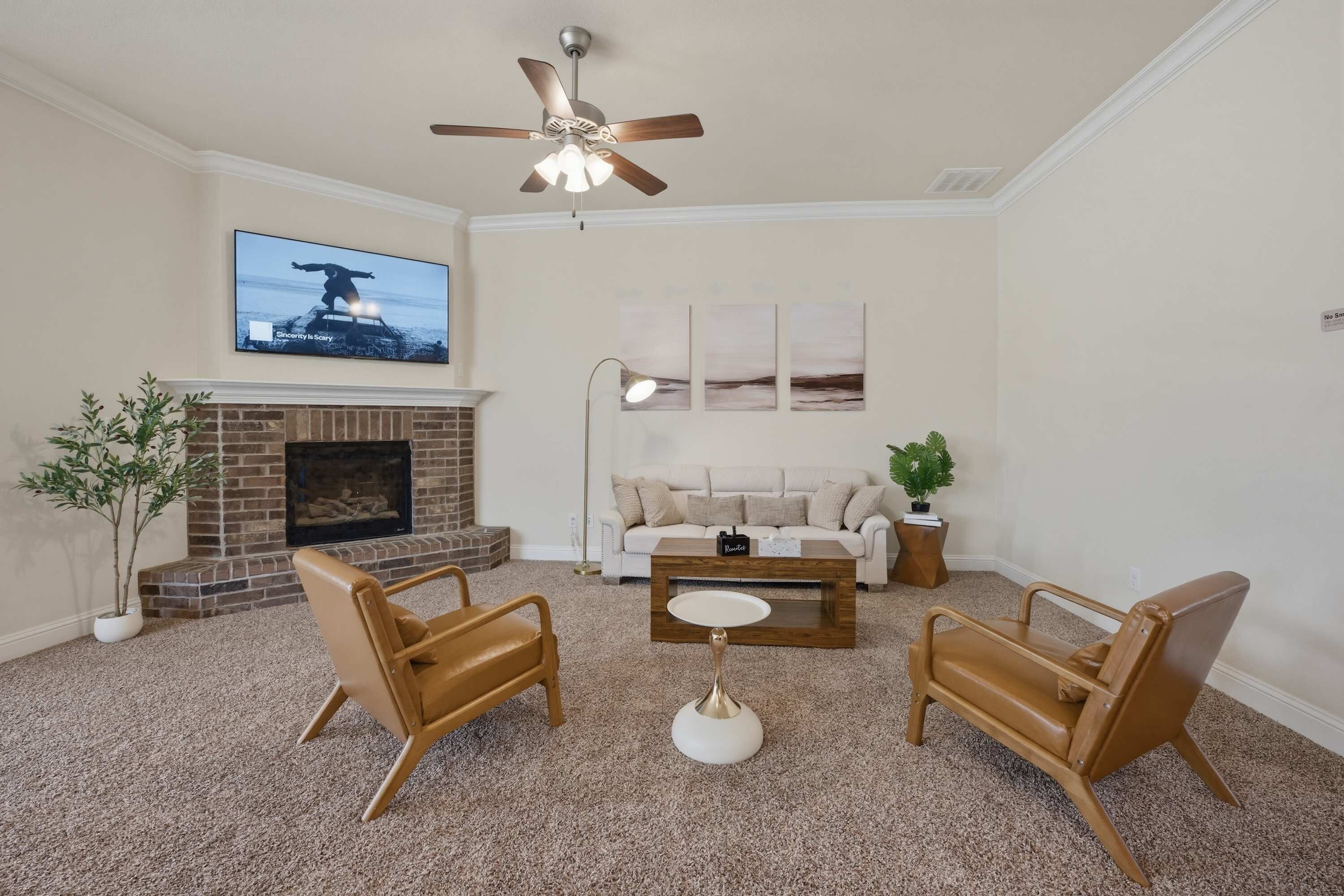 A living room with a fireplace, two brown leather chairs, a white coffee table, a ceiling fan, and a TV mounted on the wall.