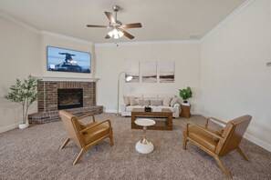 Un salon avec une cheminée, deux fauteuils en cuir marron, une table basse blanche, un ventilateur de plafond et un téléviseur fixé au mur.