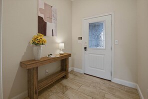 Un couloir avec une console en bois, un vase rempli de fleurs jaunes, une lampe fixée au mur et une porte dotée d’un panneau en verre.