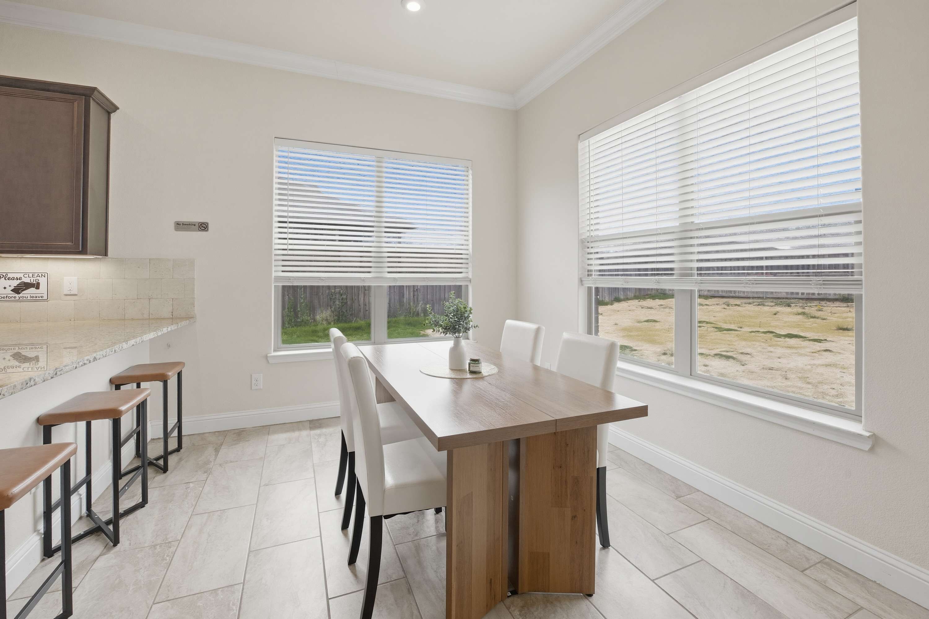 A modern kitchen with a wooden table and bar stools, large windows with blinds, and a view of the outdoors.