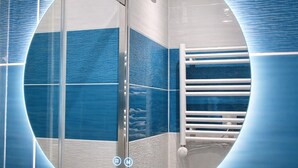 A bathroom with a circular mirror, blue tiles, a glass shower enclosure, and a white towel rack. - Blue Lagoon | Cozy Duplex • Parking • Disneyland, 10 min (Bussy-Saint-Georges)