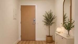 Ein Flur mit Holzboden, einem runden Teppich, einer Tür, einer Topfpflanze, einer Kommode mit Schubladen, einem Spiegel und einer Deckenleuchte.