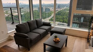 Ein modernes Wohnzimmer mit Sofa, Hocker und Couchtisch, großen Fenstern mit Stadtblick und Holzboden.