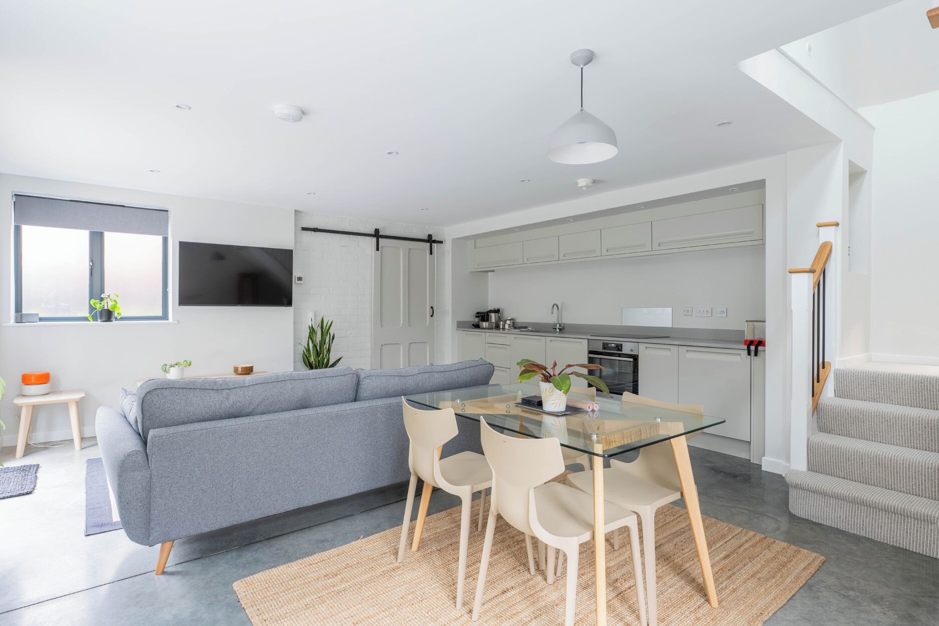 Eine moderne Küche mit weißen Schränken, einem Essbereich mit Glastisch und Stühlen, einem grauen Sofa und einem an der Wand montierten Fernseher.