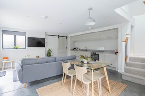 A modern kitchen with white cabinets, a dining area with a glass table and chairs, a gray sofa, and a TV mounted on the wall.