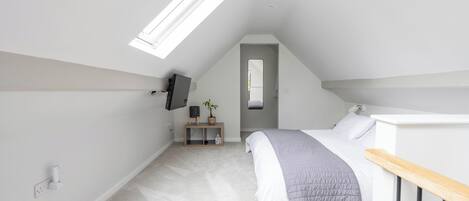 A bedroom with a sloped ceiling, a bed, a bedside table, a mirror, a TV mounted on the wall, and a skylight.