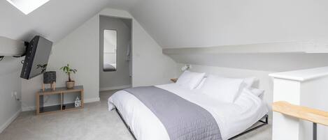 A bedroom with a sloped ceiling, a bed with white and gray bedding, a small bedside table, a mirror, a potted plant, and a desk with a chair.