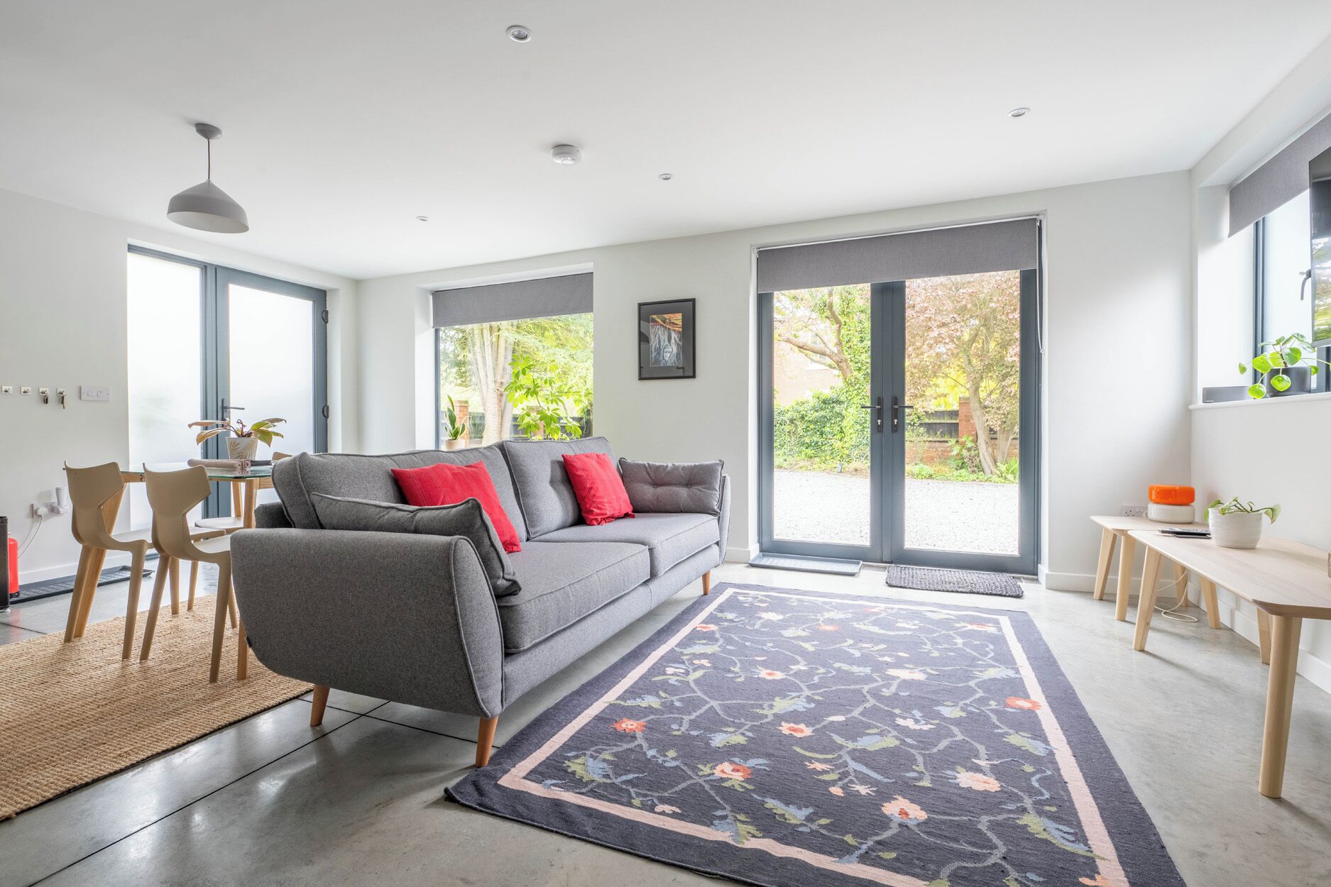 A modern living room with a grey sofa, red cushions, a wooden dining table, and a large window with a view of greenery.