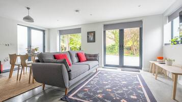 A modern living room with a grey sofa, red cushions, a wooden dining table, and a large window with a view of greenery.
