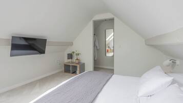 A bedroom with a bed, a TV mounted on the wall, a small wooden shelf with a plant, and a mirror reflecting a bathroom.