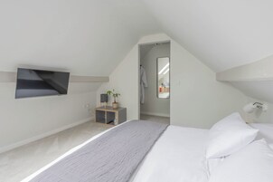 Un dormitorio con cama, televisor montado en la pared, una pequeña estantería con una planta y un espejo que refleja un baño.