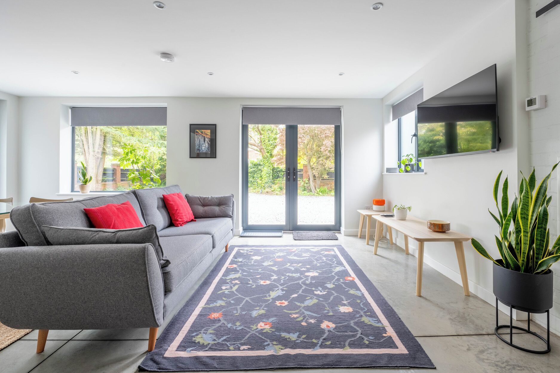 A modern living room with a grey sofa, red cushions, a floral rug, and a large window with a view of greenery.
