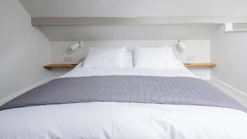 Una cama bien tendida con ropa blanca y una manta gris, dos mesitas de noche con lámparas, y un estante de madera en la pared.