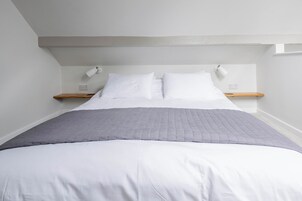 Una cama bien tendida con ropa blanca y una manta gris, dos mesitas de noche con lámparas, y un estante de madera en la pared.