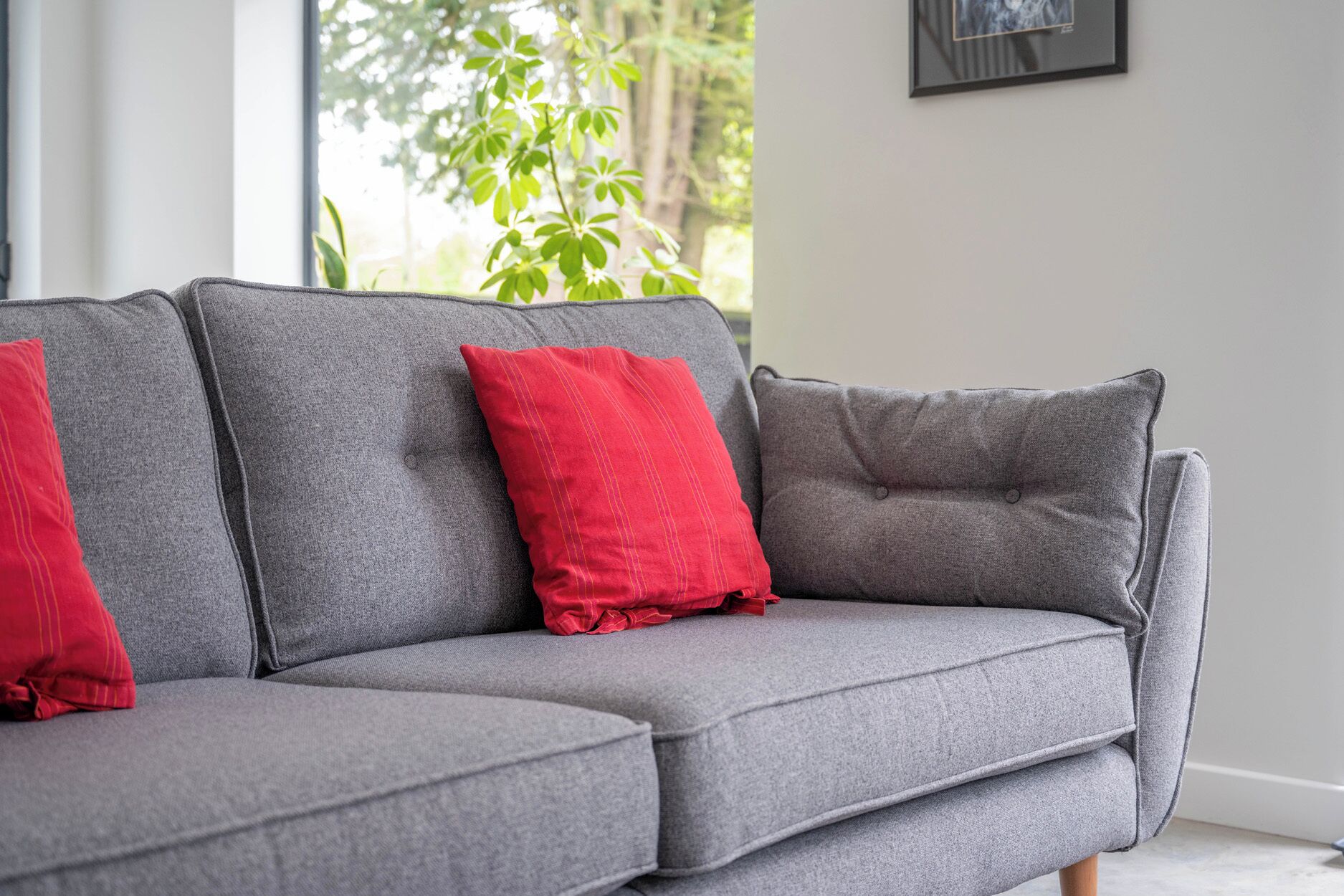 A grey sectional sofa with red cushions in a room with a window and a framed picture on the wall.