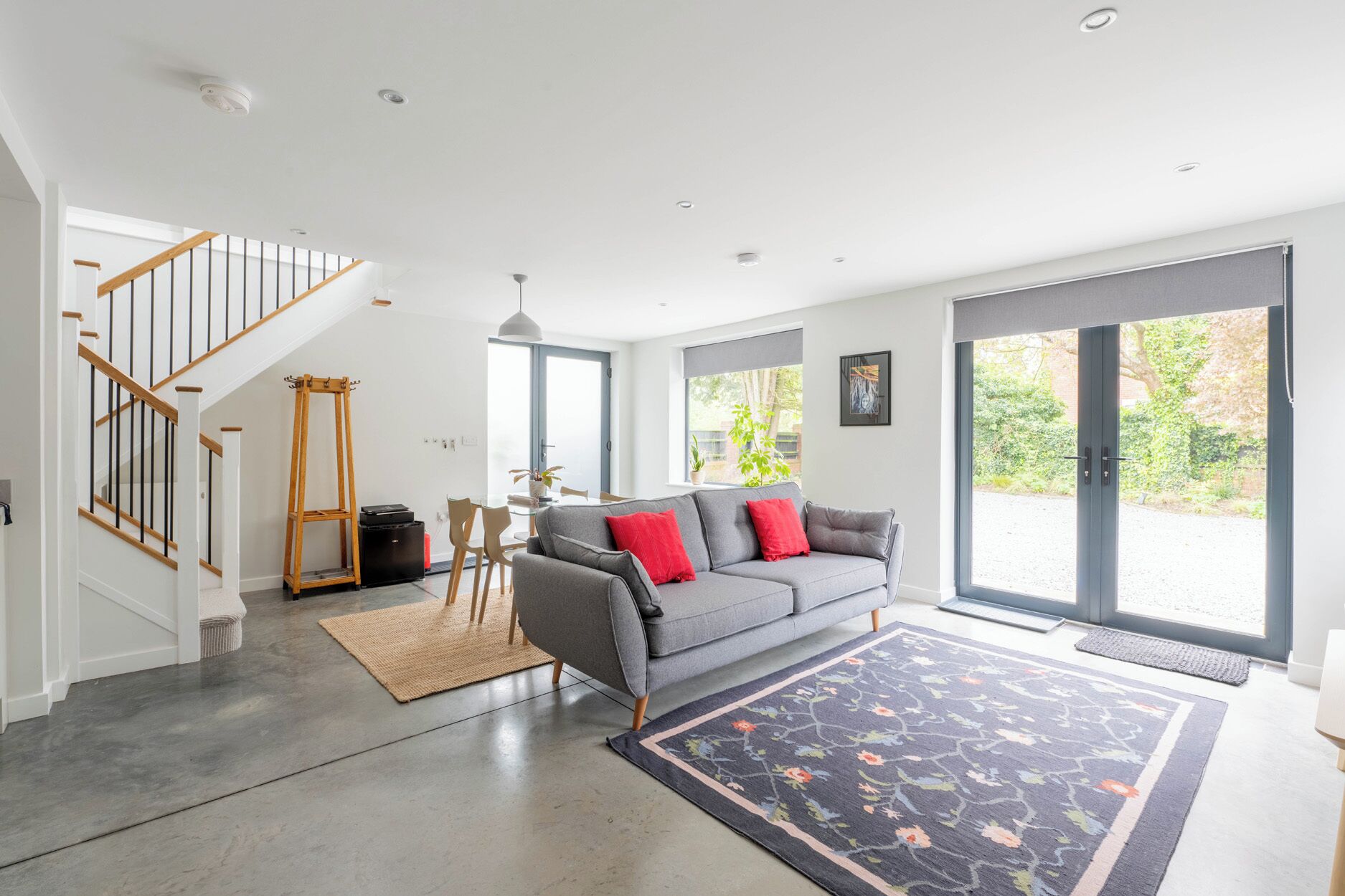 Een moderne woonkamer met een grijze bank, rode kussens, een houten trap en een groot raam.
