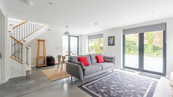 Een moderne woonkamer met een grijze bank, rode kussens, een houten trap en een groot raam.