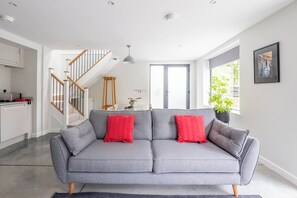 A modern living room with a grey sofa, red cushions, a staircase, and a large window.