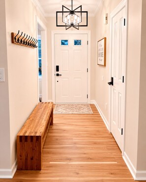 A hallway with a wooden bench, a chandelier, a door with a picture frame, and a wall-mounted coat rack.