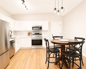 A modern kitchen with white cabinets, stainless steel appliances, a wooden dining table with black chairs, and pendant lighting.