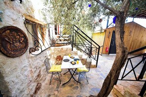 Un espacio de estar al aire libre con paredes de piedra, una mesa, sillas, una escalera metálica y un árbol.