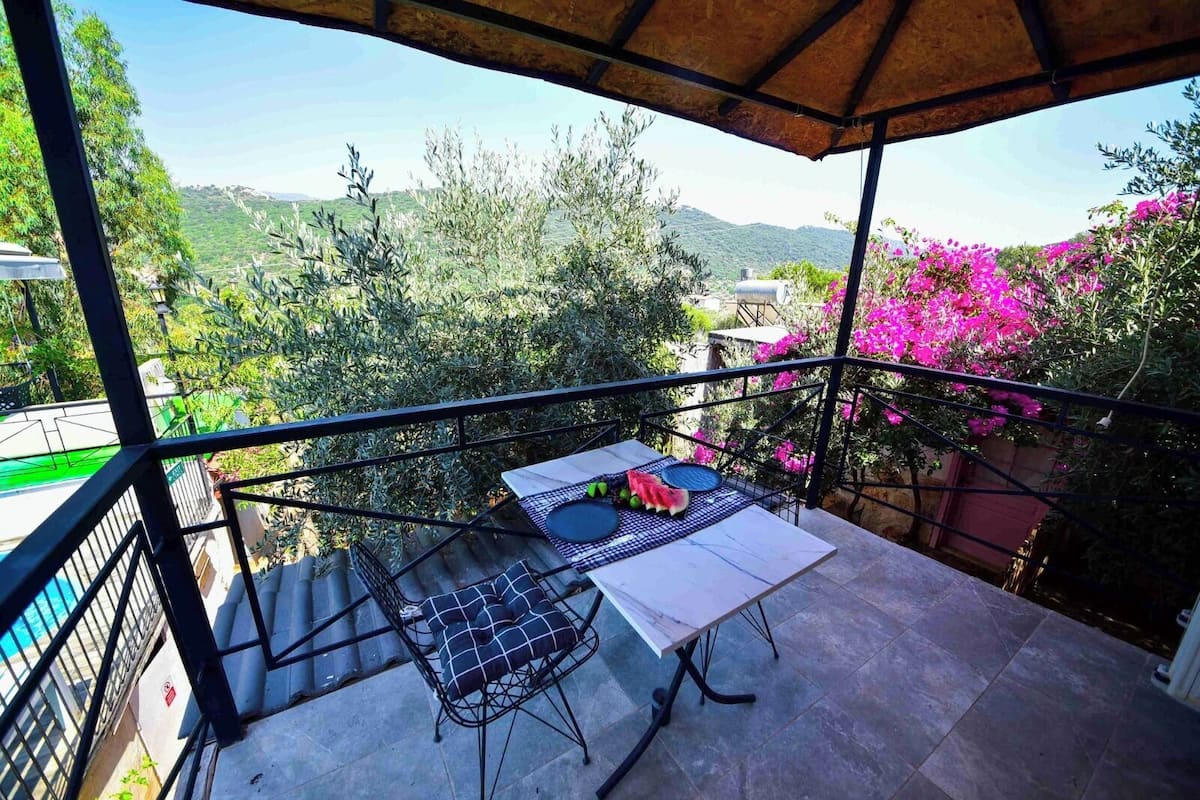 A balcony with a table set for two, overlooking a mountainous landscape with greenery and pink flowers.