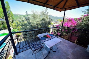 Un balcón con una mesa dispuesta para dos personas, con vistas a un paisaje montañoso con vegetación y flores rosadas.