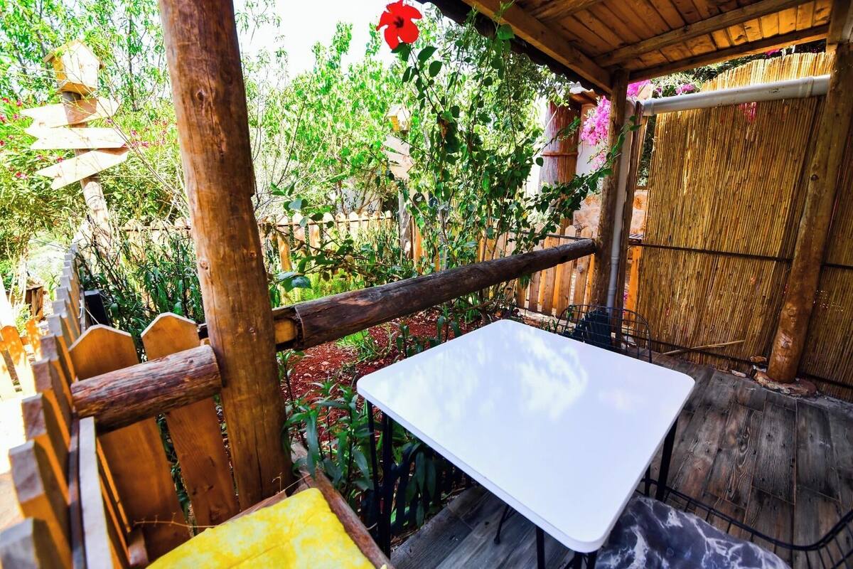 A wooden deck with a white table and a yellow cushion, surrounded by a bamboo fence and lush greenery.