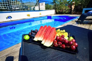 Mesa junto a la piscina con una bandeja de frutas, piscina y sillas de descanso.