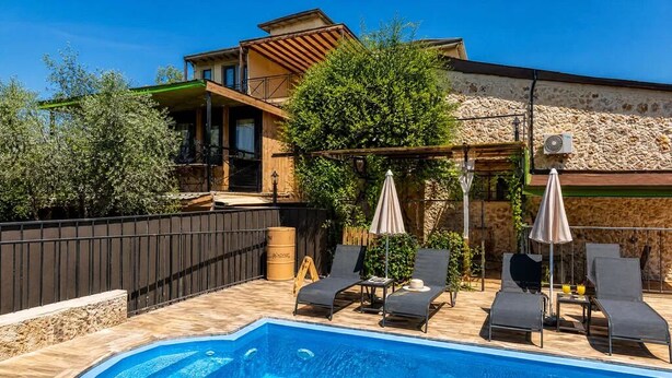 A poolside area with lounge chairs and umbrellas, a stone wall, and a building with a balcony.