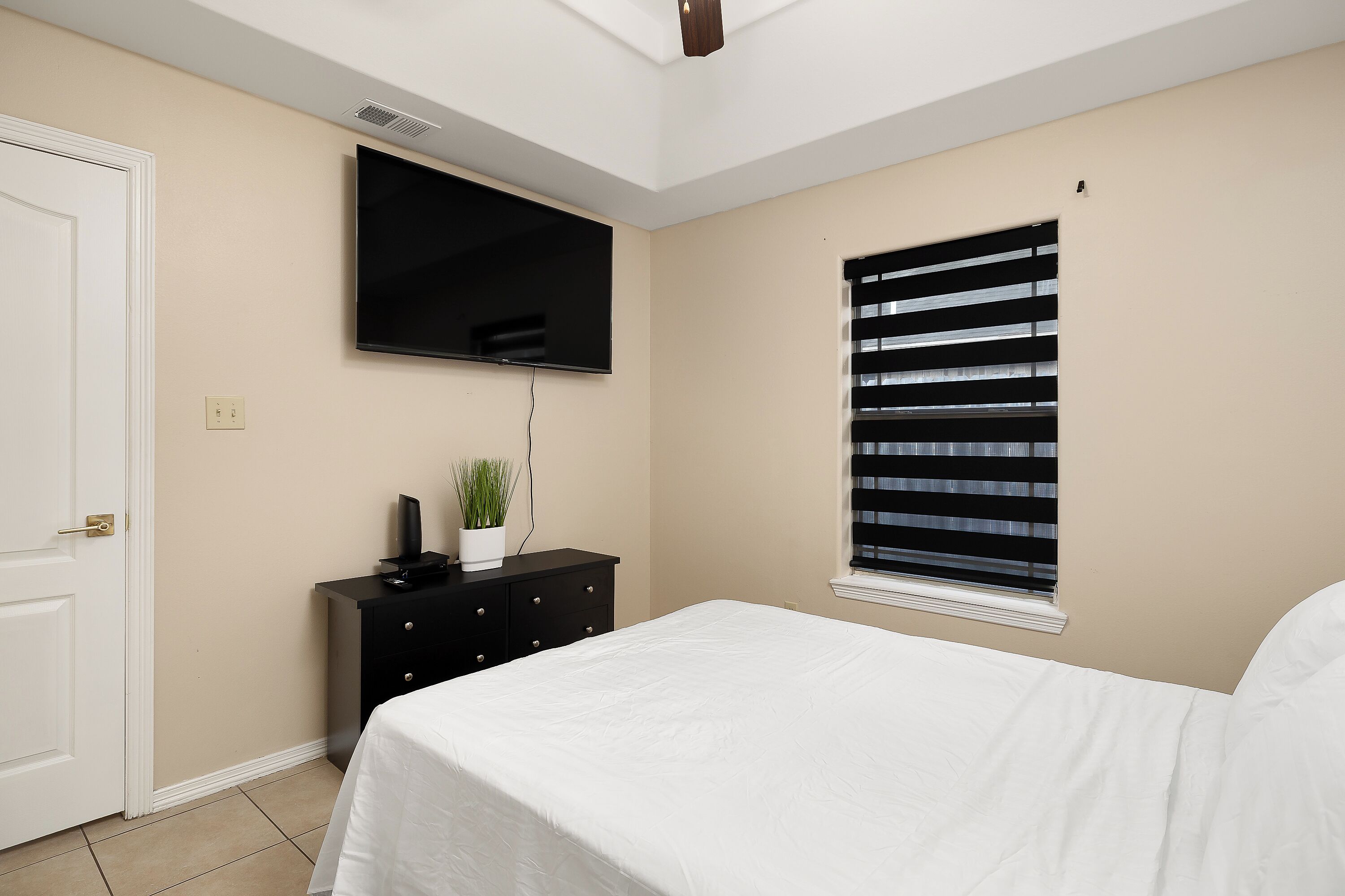 A bedroom with a bed, a TV mounted on the wall, a window with blinds, and a door.
