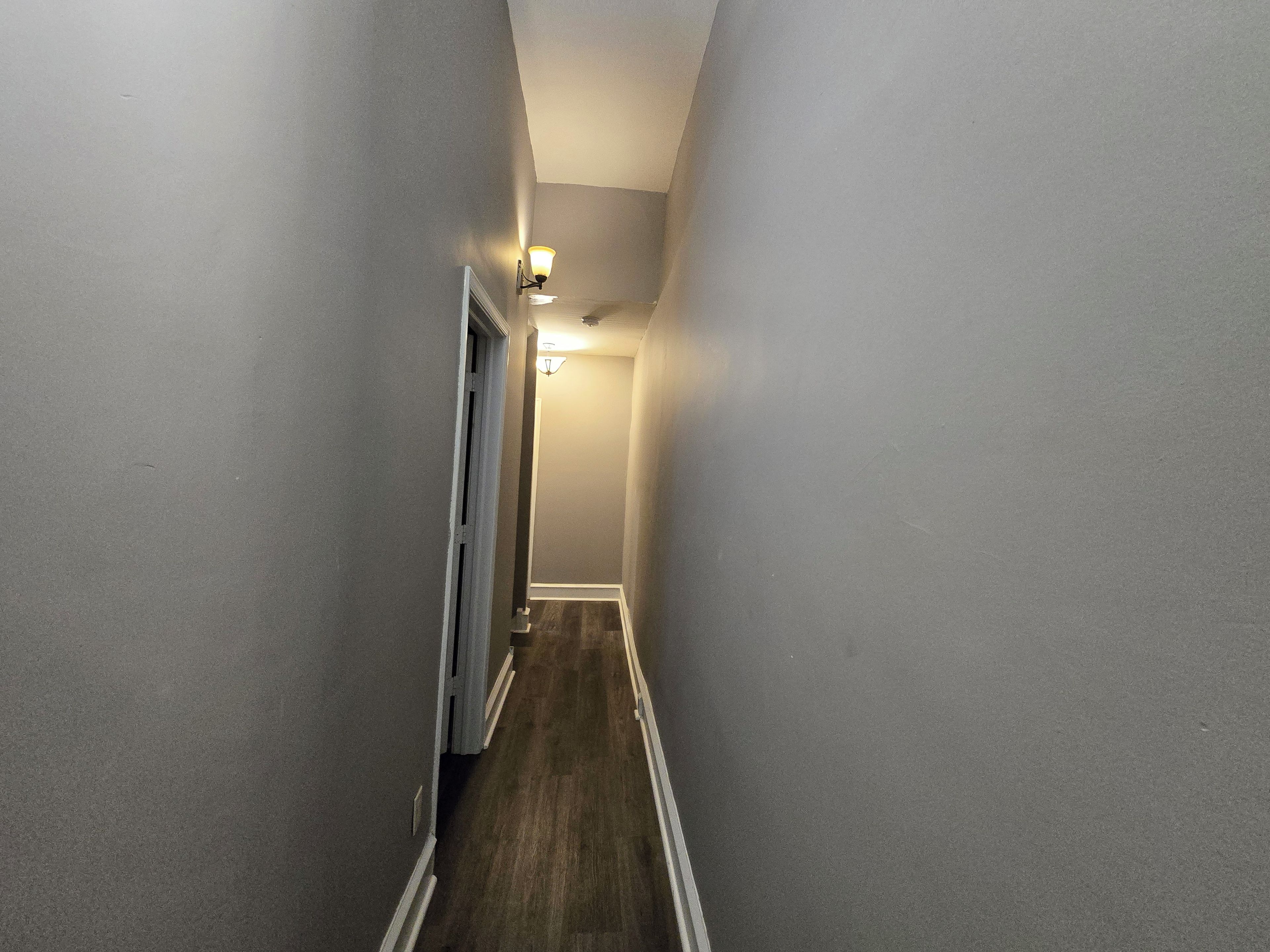 A narrow hallway with a wooden floor and white trim, leading to a door at the end.