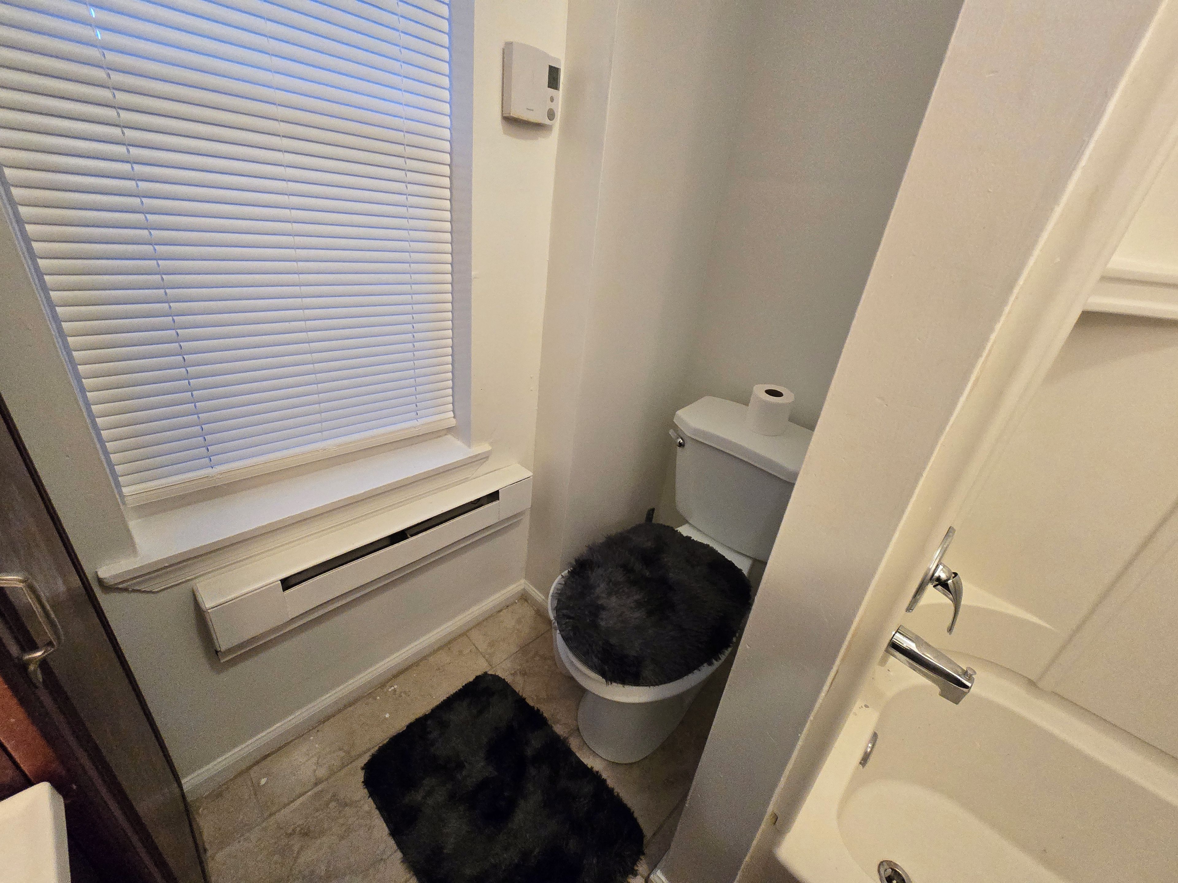 A bathroom with a toilet, a window with blinds, a black rug, and a white wall-mounted heater.