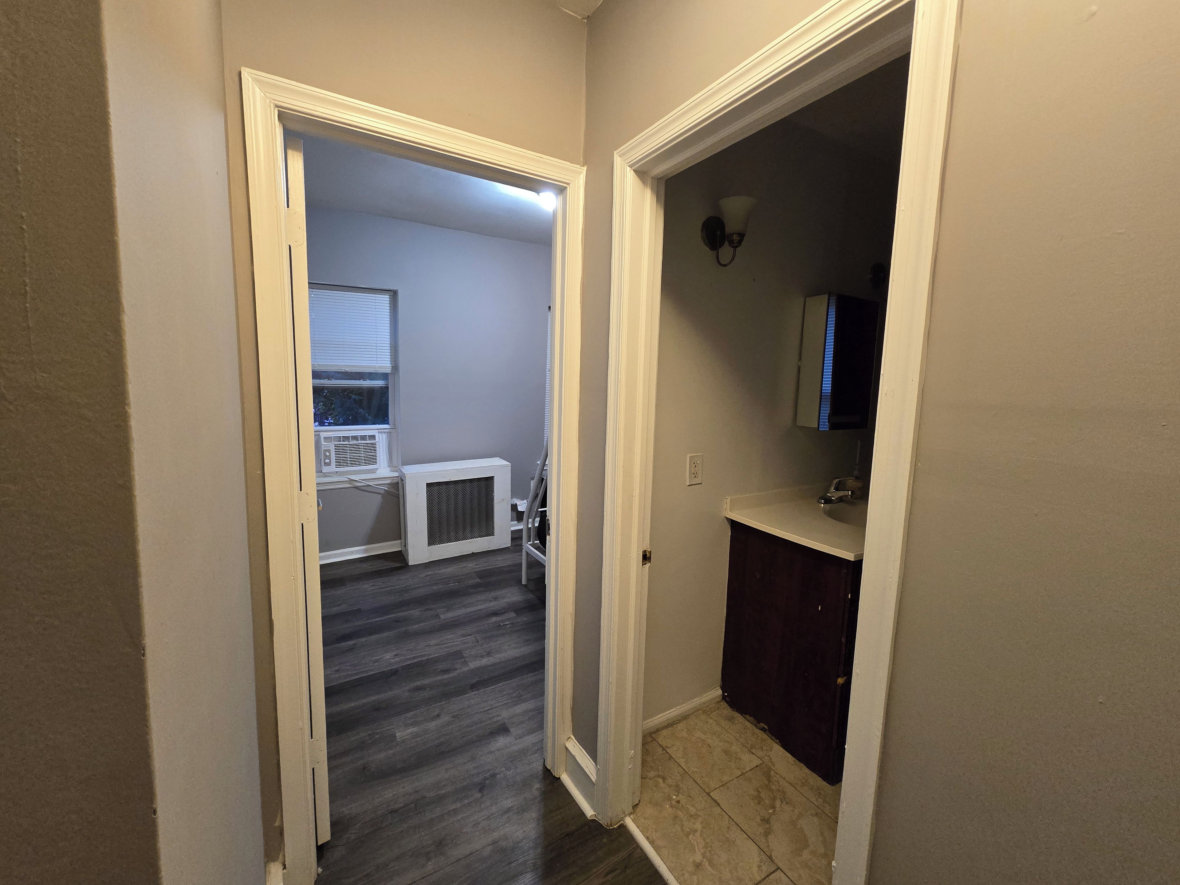 A hallway with a dark wood floor, a white air conditioning unit, and a bathroom with a dark cabinet and a white sink.