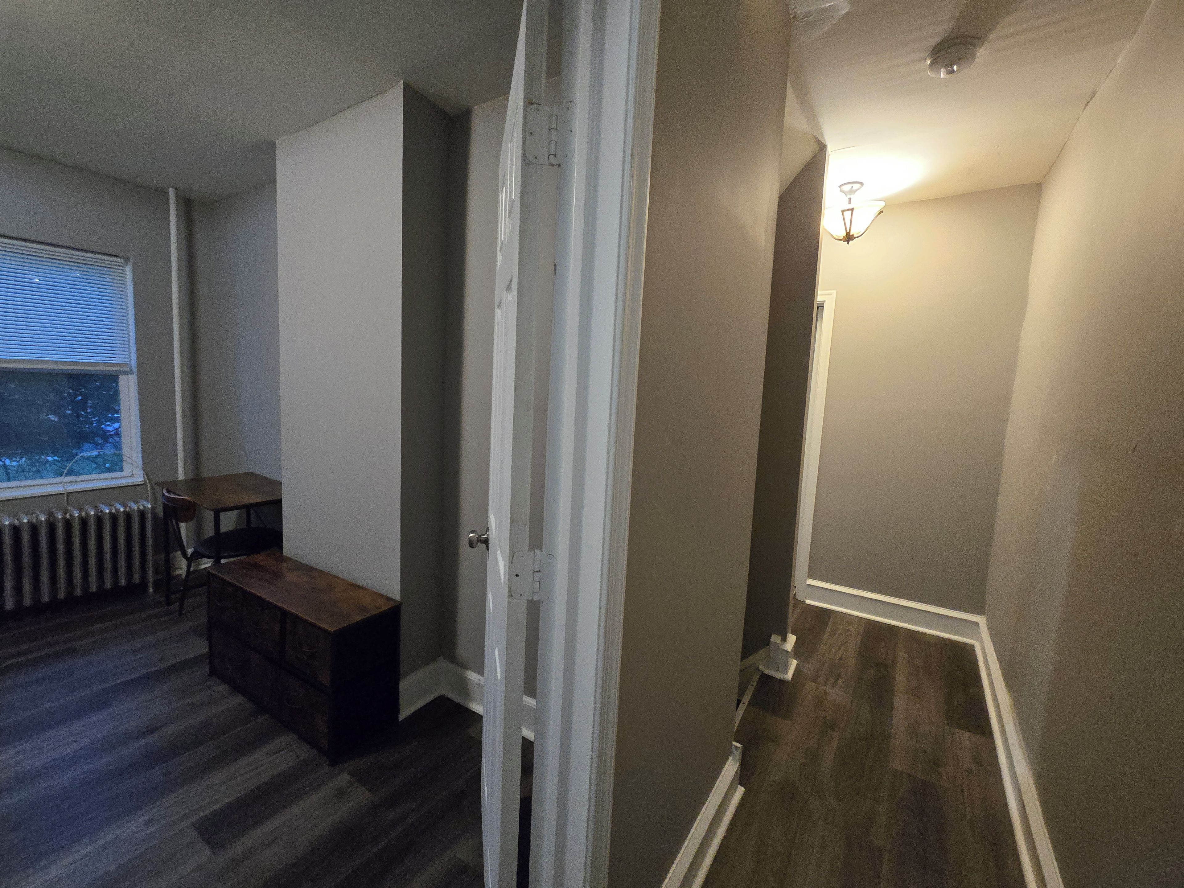 A room with a wooden floor, a radiator, a window with blinds, a small table, and a bench.