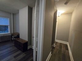 Una habitación con piso de madera, un radiador, una ventana con persianas, una mesa pequeña y un banco.