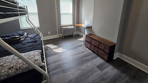 Una habitación con literas, un radiador, un banco de madera y una mesa con lámpara.