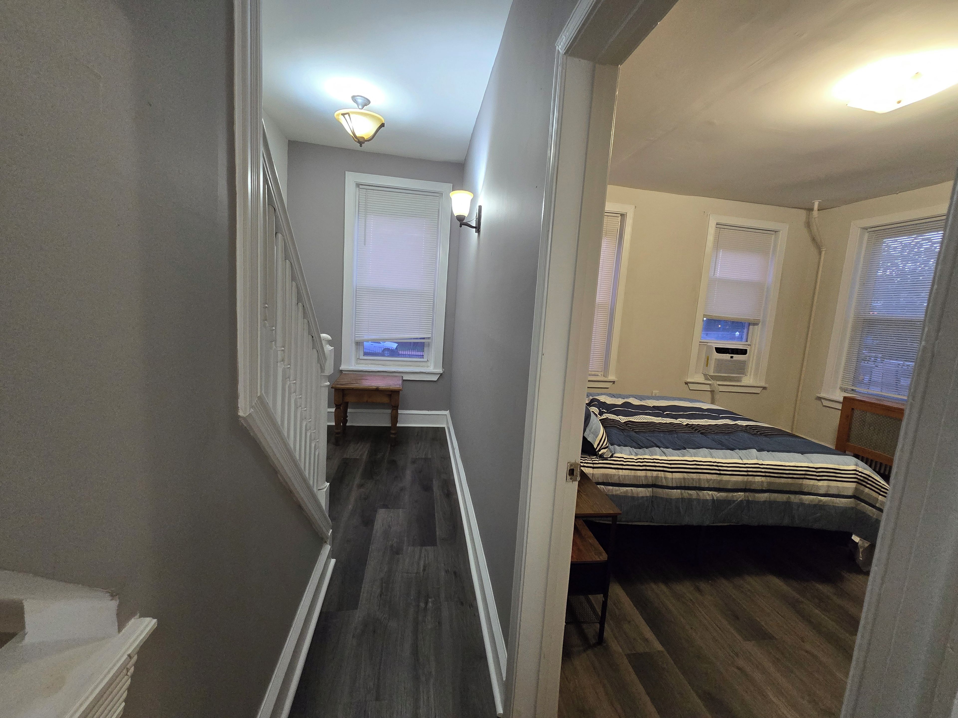 A narrow hallway with a staircase leading to a bedroom. The room has a bed with a striped bedspread, a small table, and a window with blinds.