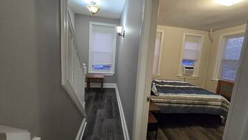 A narrow hallway with a staircase leading to a bedroom. The room has a bed with a striped bedspread, a small table, and a window with blinds.